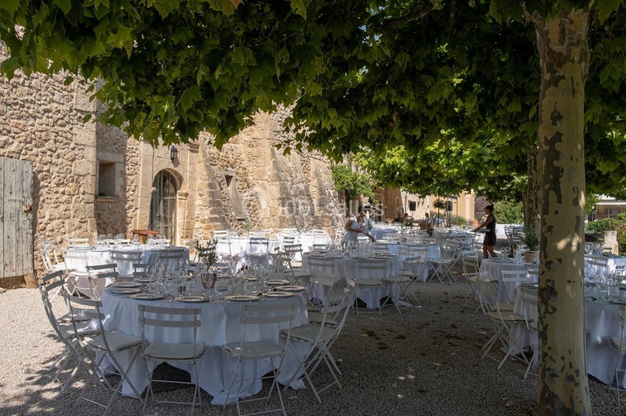 Tables rondes dressées avec nappes blanches sous des arbres, devant un bâtiment en pierre dans une cour extérieure.