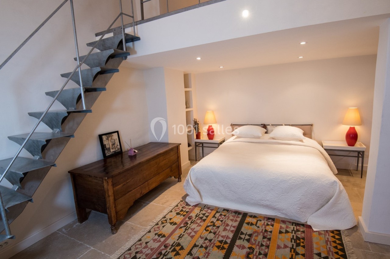 Chambre avec lit double, tapis coloré, commode en bois, lampes rouges et escalier menant à une mezzanine.