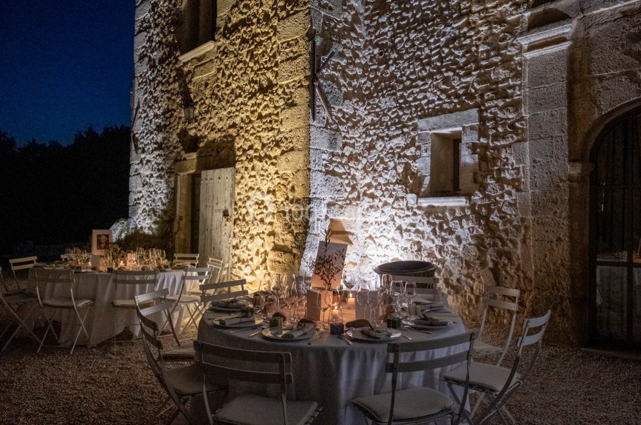 Tables rondes dressées pour un dîner en extérieur, éclairées par des bougies, devant un mur en pierre éclairé.
