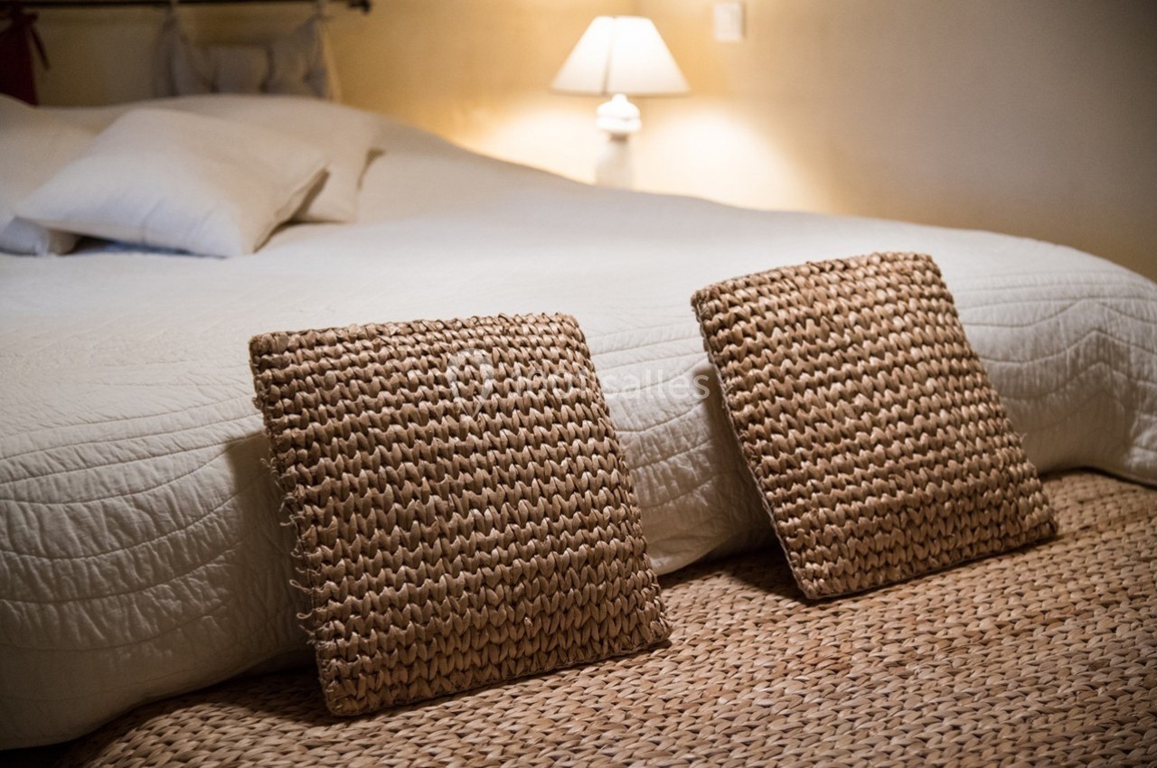 Lit avec une couverture blanche, deux coussins en osier au sol et une lampe allumée en arrière-plan.