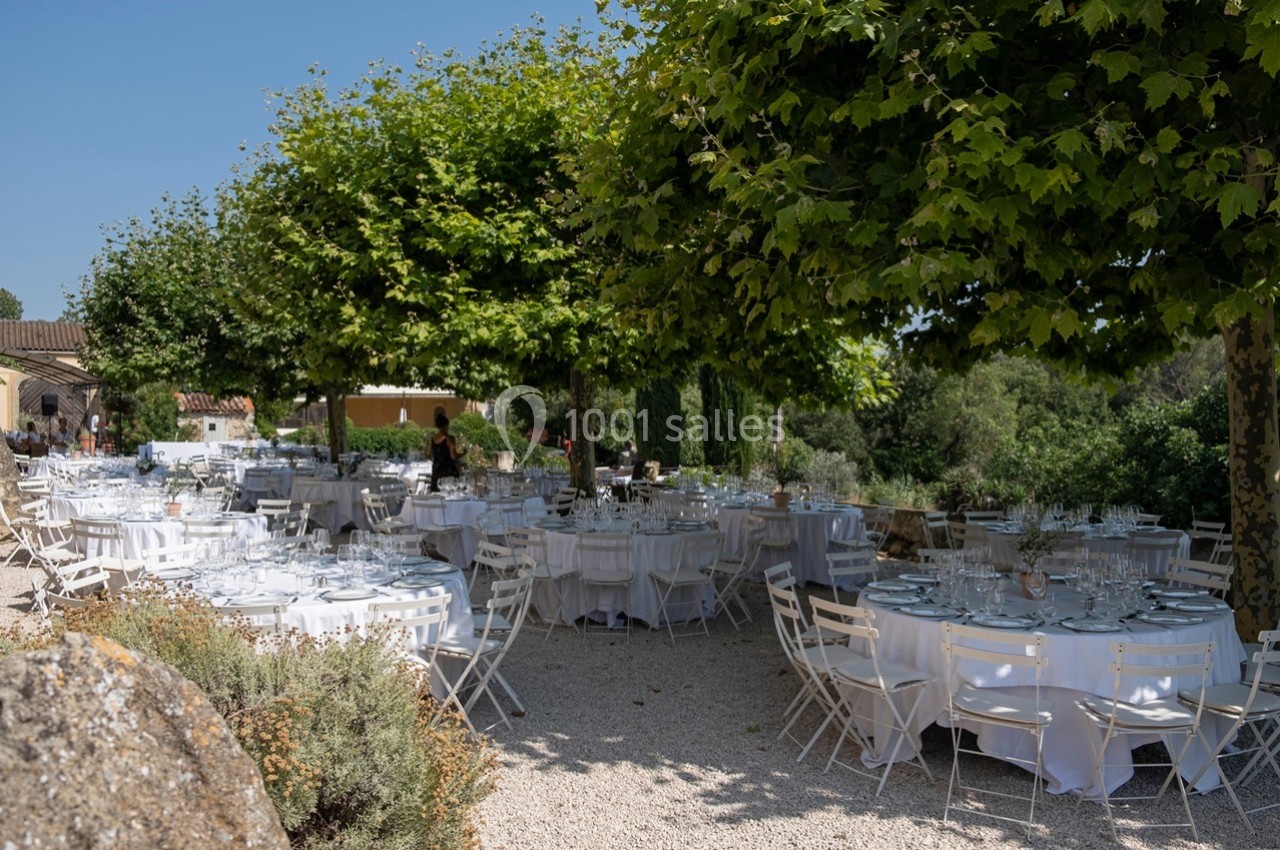 Tables rondes dressées avec nappes blanches sous des arbres dans un espace extérieur ensoleillé.