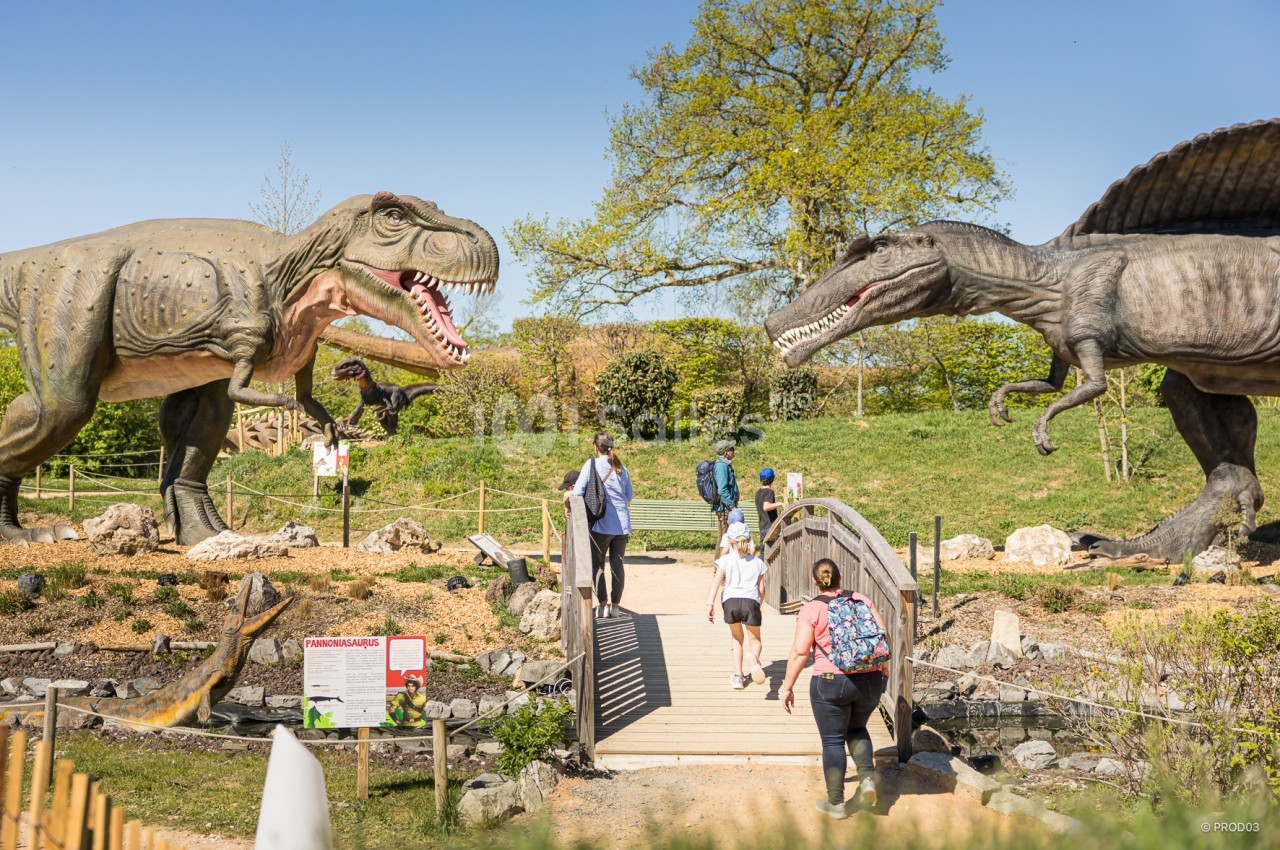 Des visiteurs traversent un pont dans un parc avec des statues grandeur nature de dinosaures sous un ciel dégagé.
