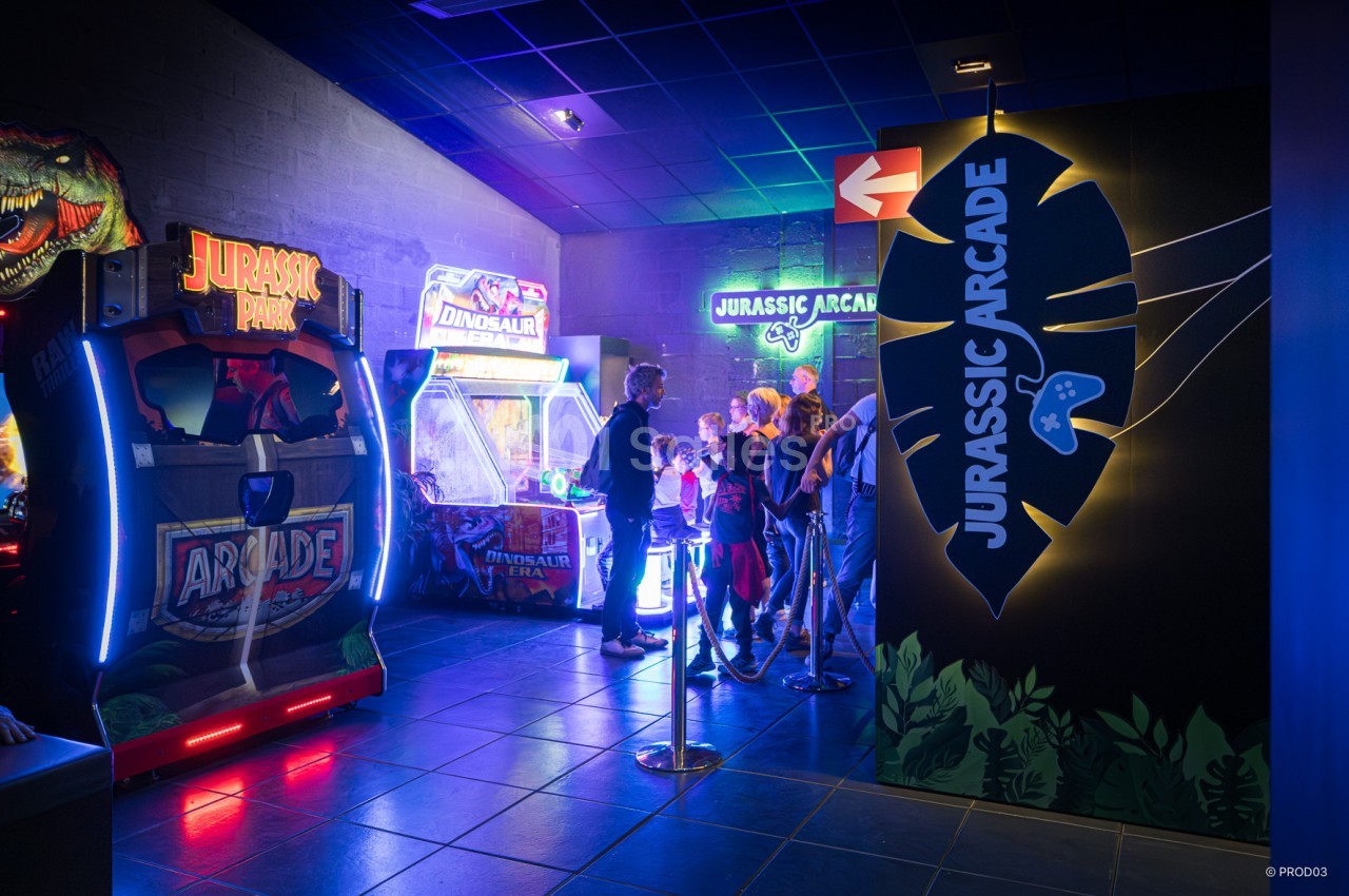 Des personnes font la queue devant des bornes d'arcade dans une salle de jeux à thème Jurassic.