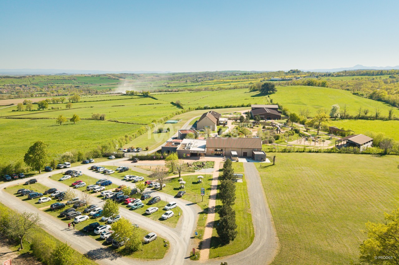 Vue aérienne d'un complexe rural avec bâtiments, parking rempli de voitures et paysage verdoyant environnant.