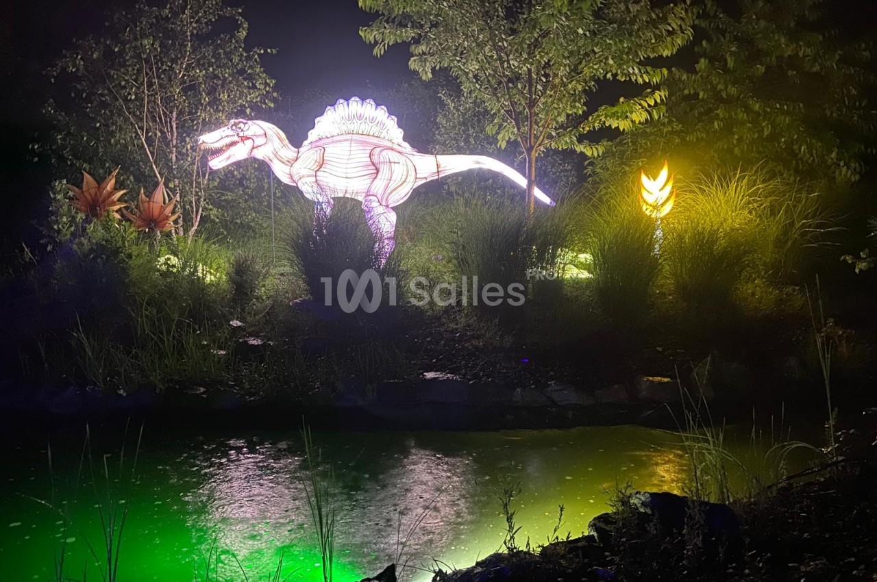 Reproduction lumineuse d'un dinosaure près d'un étang éclairé, entouré de végétation et de lumières colorées.