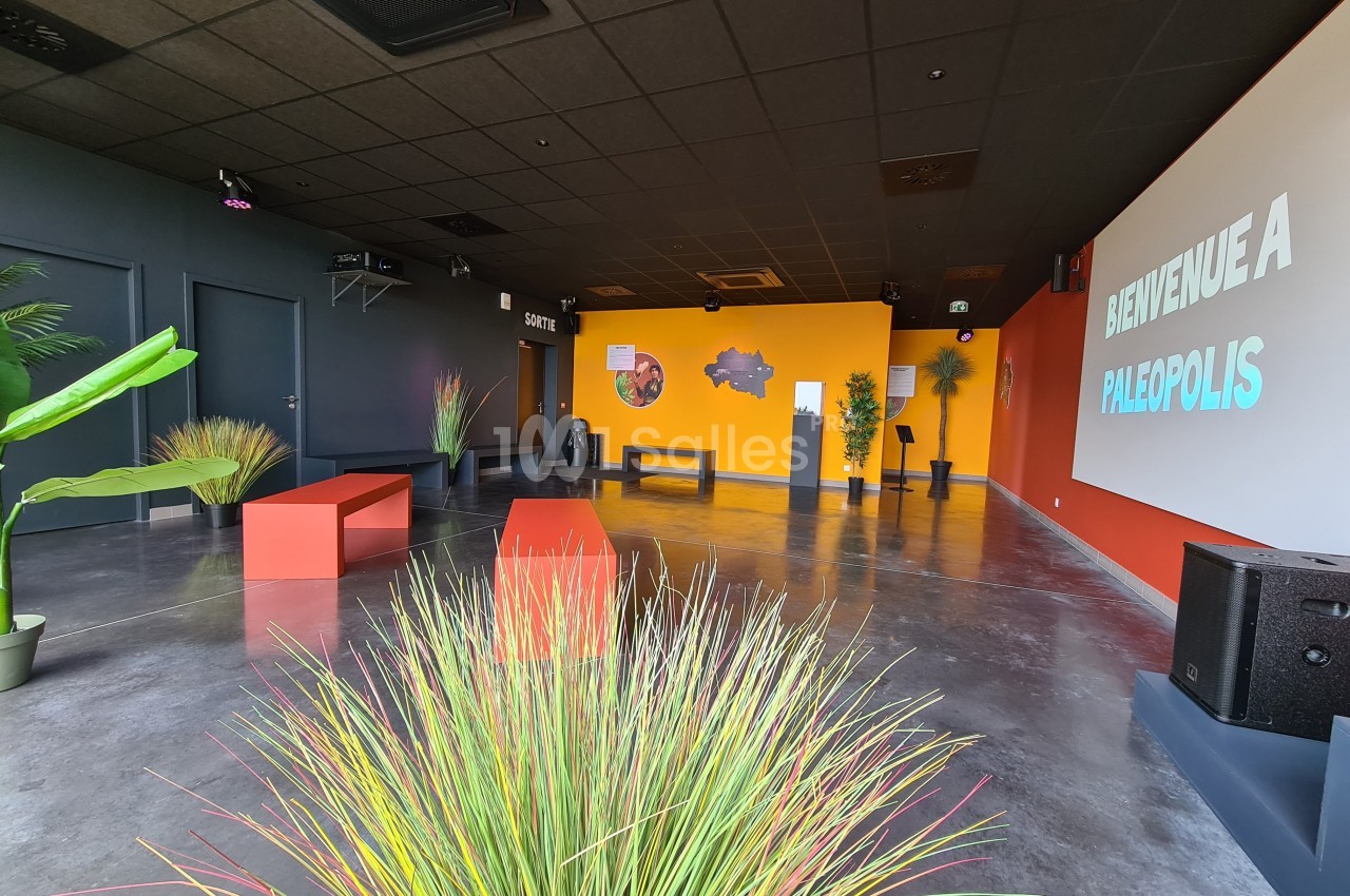 Salle d'accueil moderne avec bancs colorés, plantes décoratives et un écran affichant ’Bienvenue à Paléopolis’.