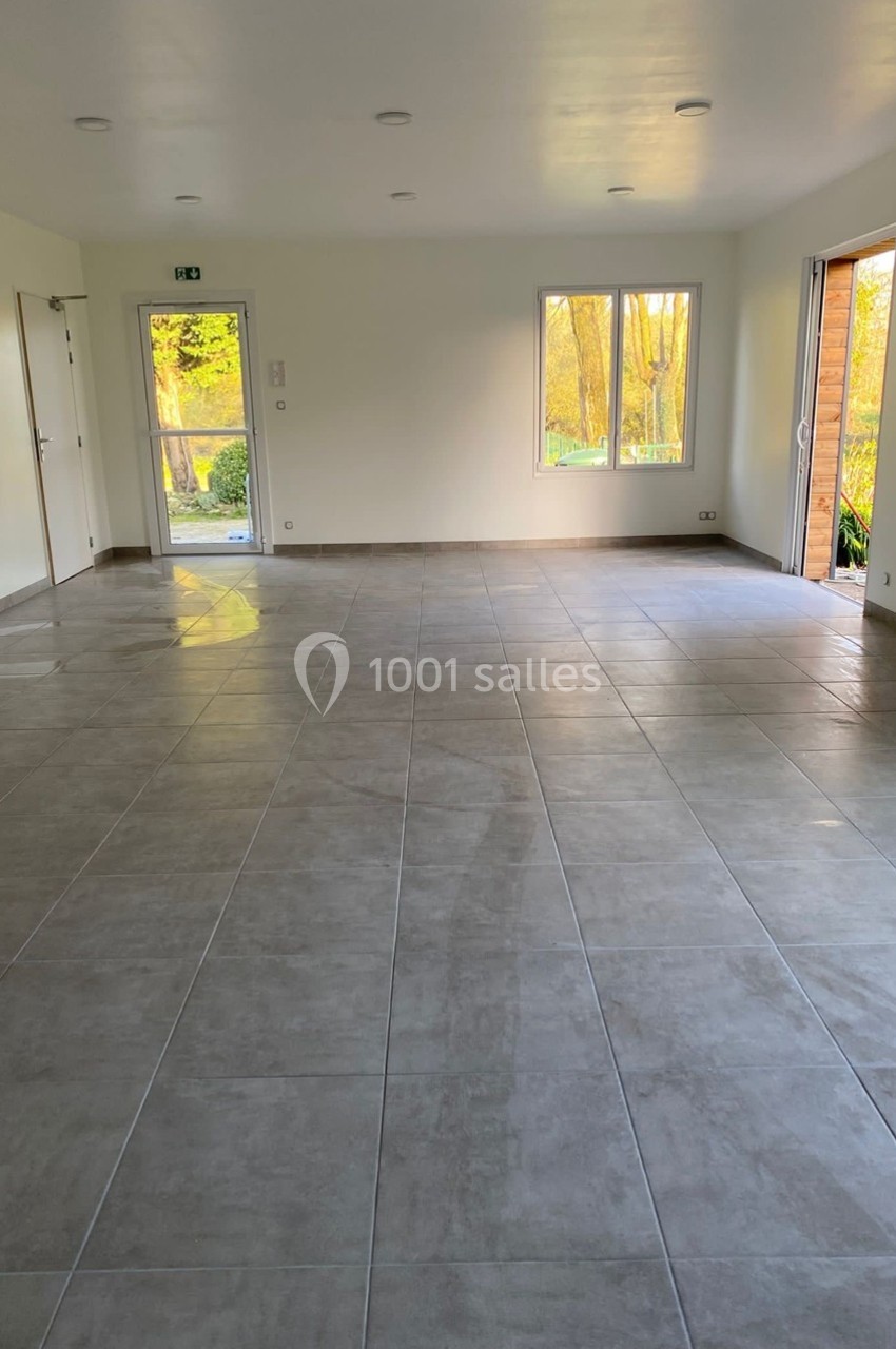 Salle vide avec carrelage gris, murs blancs, fenêtres laissant entrer la lumière naturelle et porte ouverte sur l'extérieur.