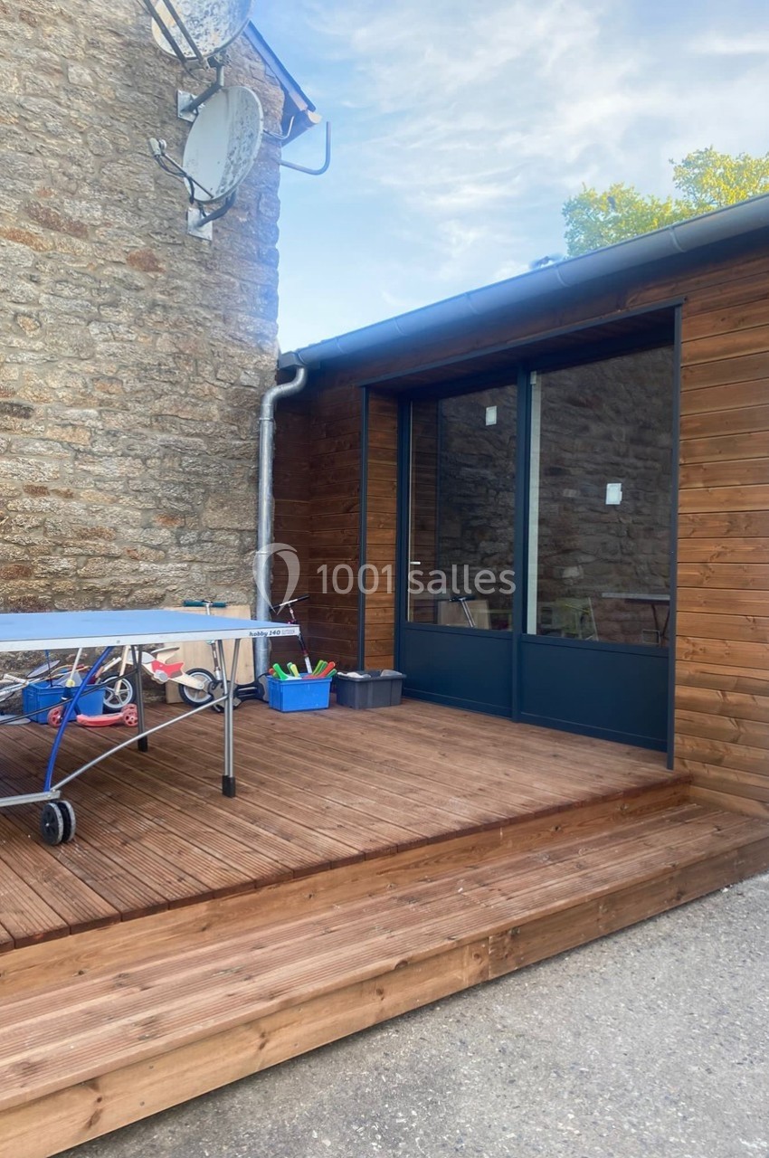 Terrasse en bois avec une table de ping-pong, jouets dans un bac et façade en pierre et bois d'une maison.
