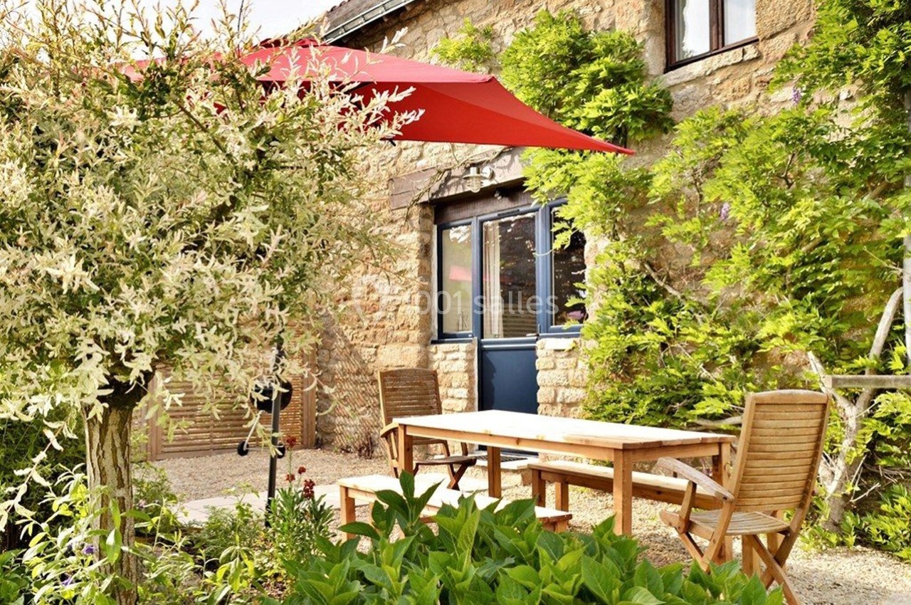 Table en bois et chaises dans un jardin fleuri, avec un parasol rouge et une façade en pierre en arrière-plan.