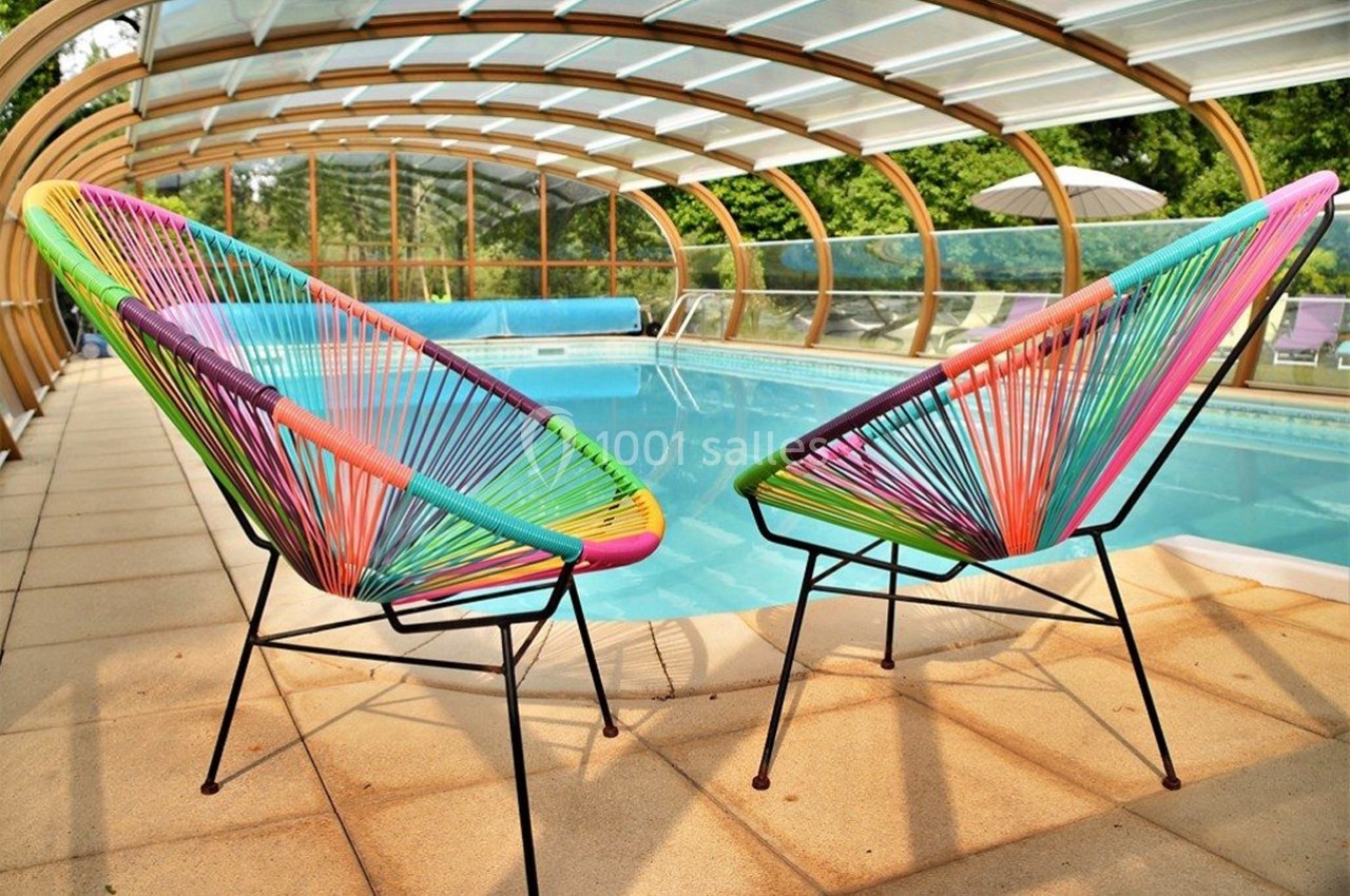 Deux fauteuils colorés en corde face à une piscine couverte par une structure transparente, entourée de verdure.