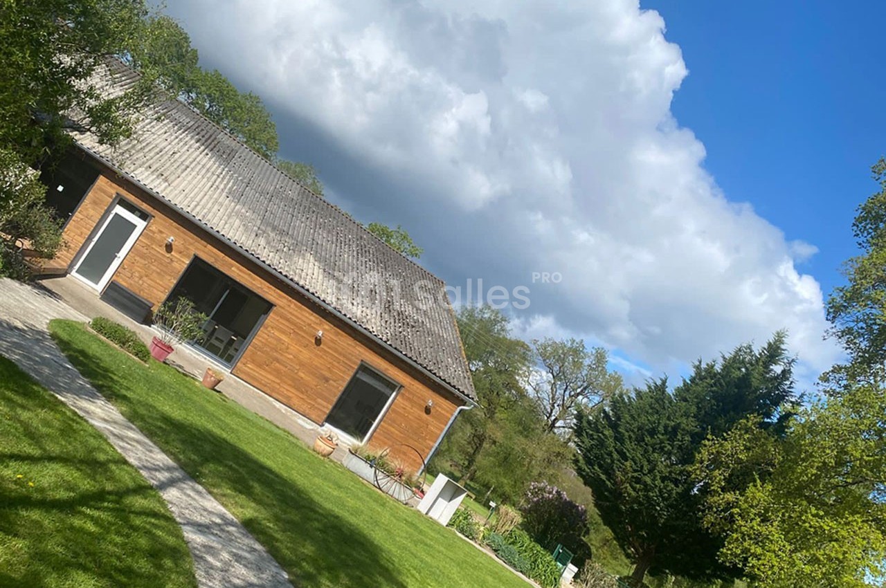 Maison en bois avec toit en tuiles, entourée d'un jardin verdoyant sous un ciel partiellement nuageux.