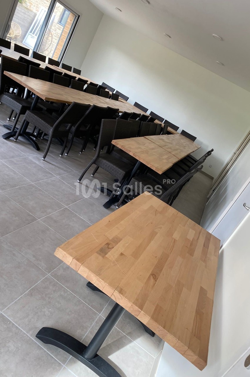Salle lumineuse avec plusieurs tables en bois clair et chaises noires, disposées en rangées sur un sol carrelé.