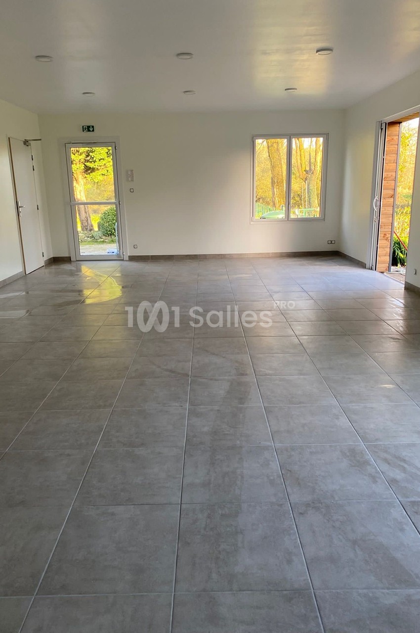 Salle vide avec carrelage gris, murs blancs, fenêtres laissant entrer la lumière naturelle et porte ouverte sur l'extérieur.