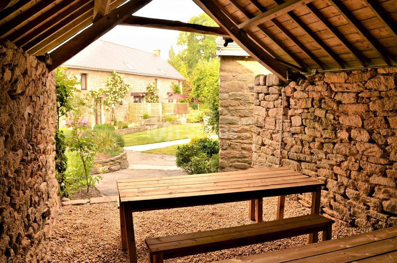Vue depuis un abri en pierre et bois sur une table en bois, donnant sur un jardin et une maison en arrière-plan.