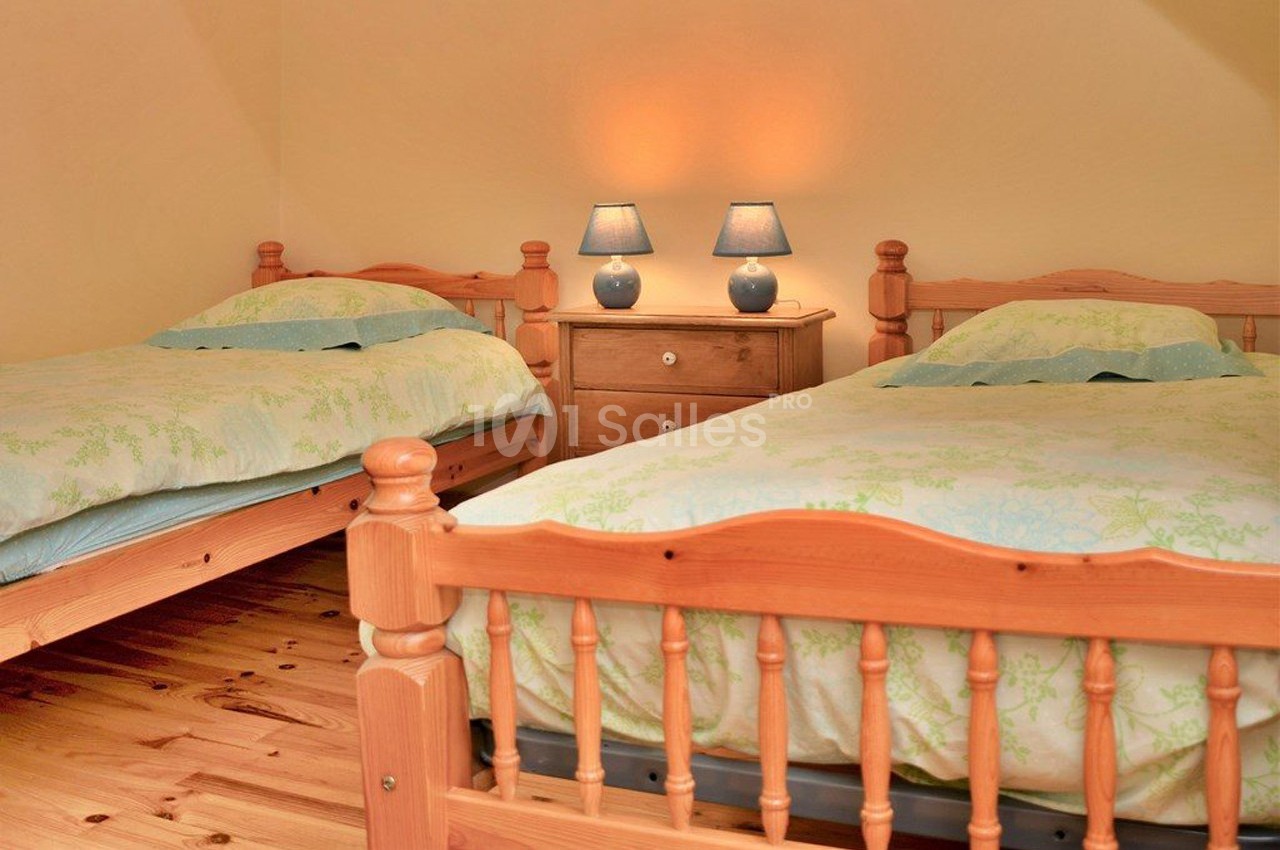 Deux lits simples en bois avec une table de chevet et deux lampes allumées dans une chambre aux tons chaleureux.