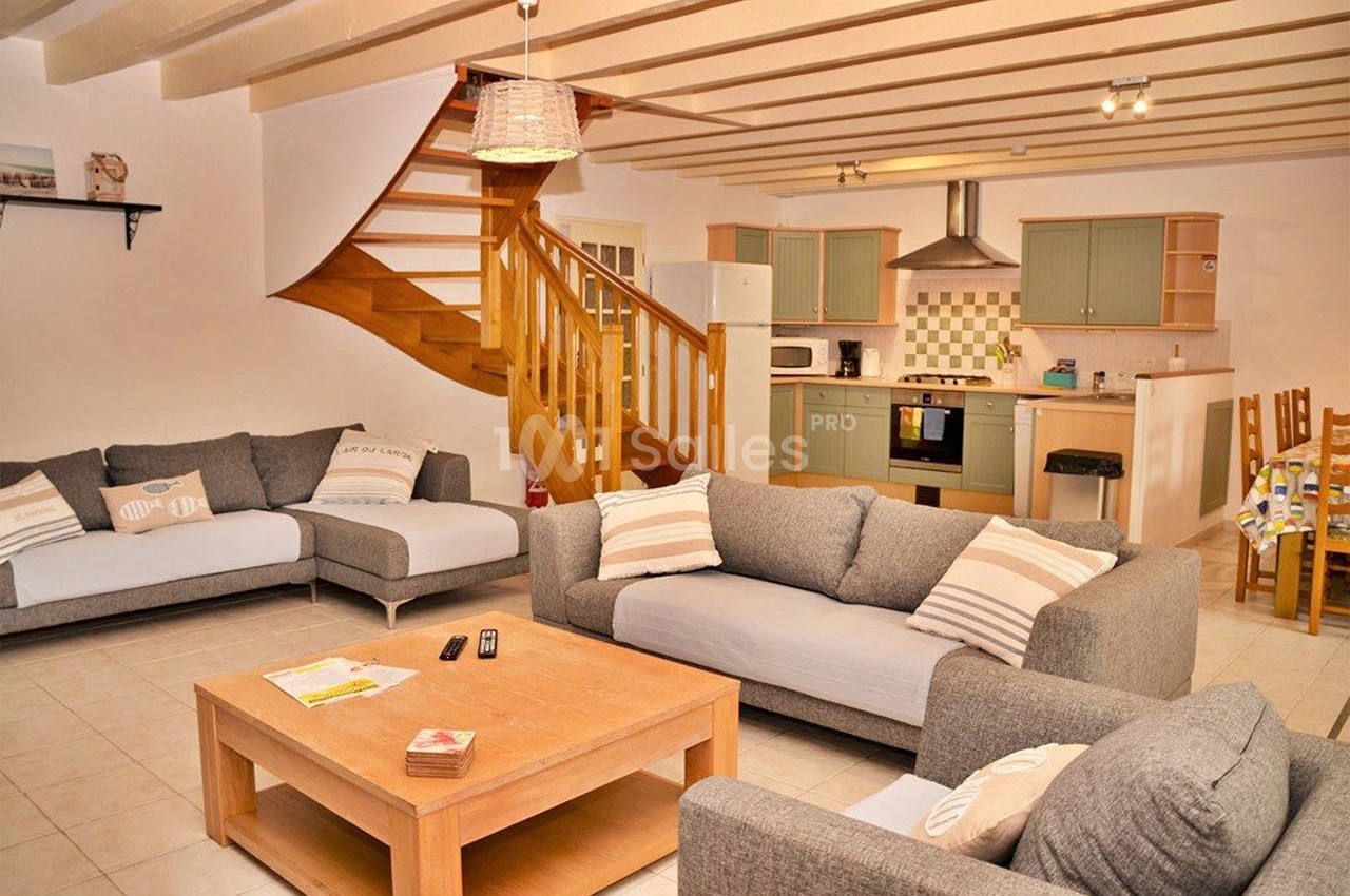 Salon lumineux avec canapés gris, table basse en bois, escalier en bois et cuisine ouverte équipée en arrière-plan.