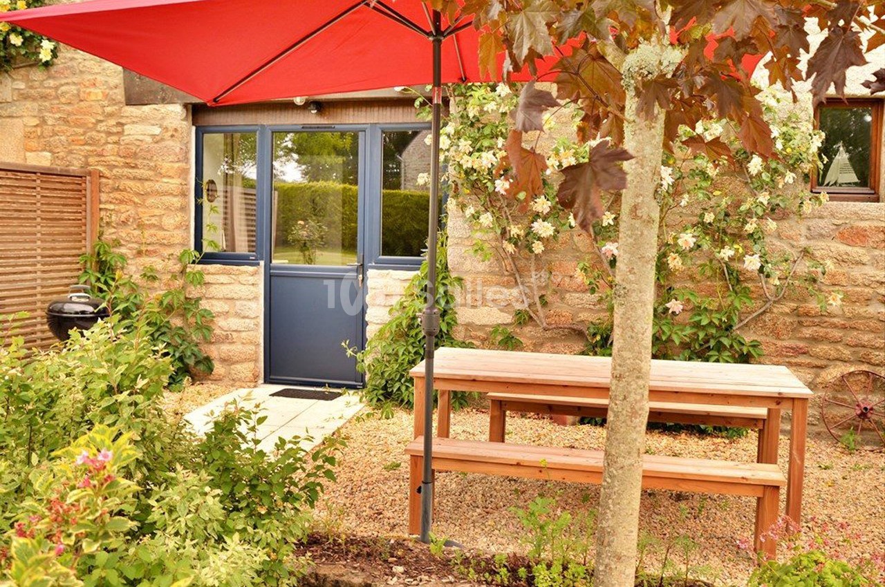 Cour extérieure avec table en bois, parasol rouge, mur en pierre, porte bleue et végétation fleurie.