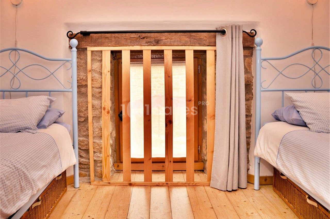 Deux lits simples avec draps bleus encadrent une petite porte en bois et pierre, partiellement couverte par un rideau beige.