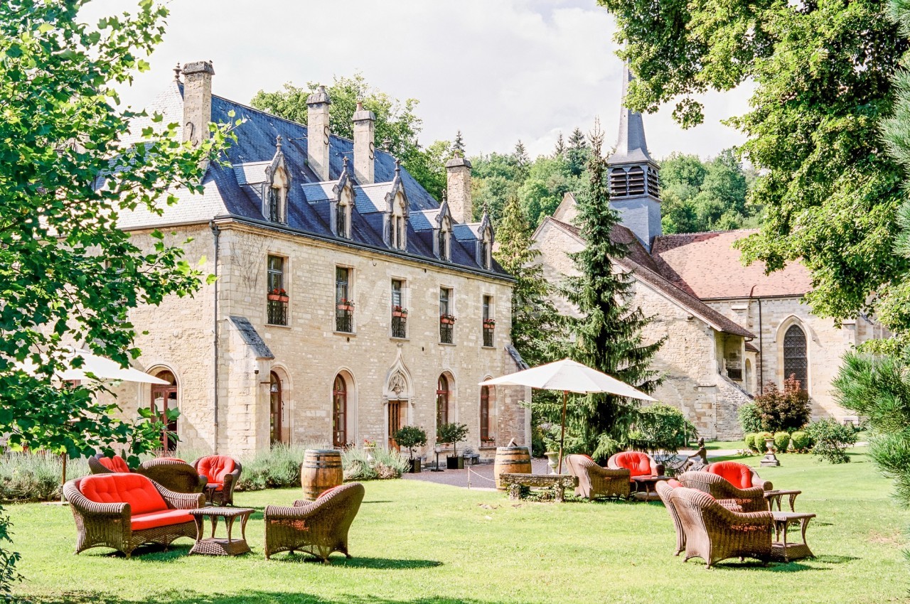 Jardin avec fauteuils en osier et parasols devant un bâtiment en pierre et une église dans un cadre verdoyant.