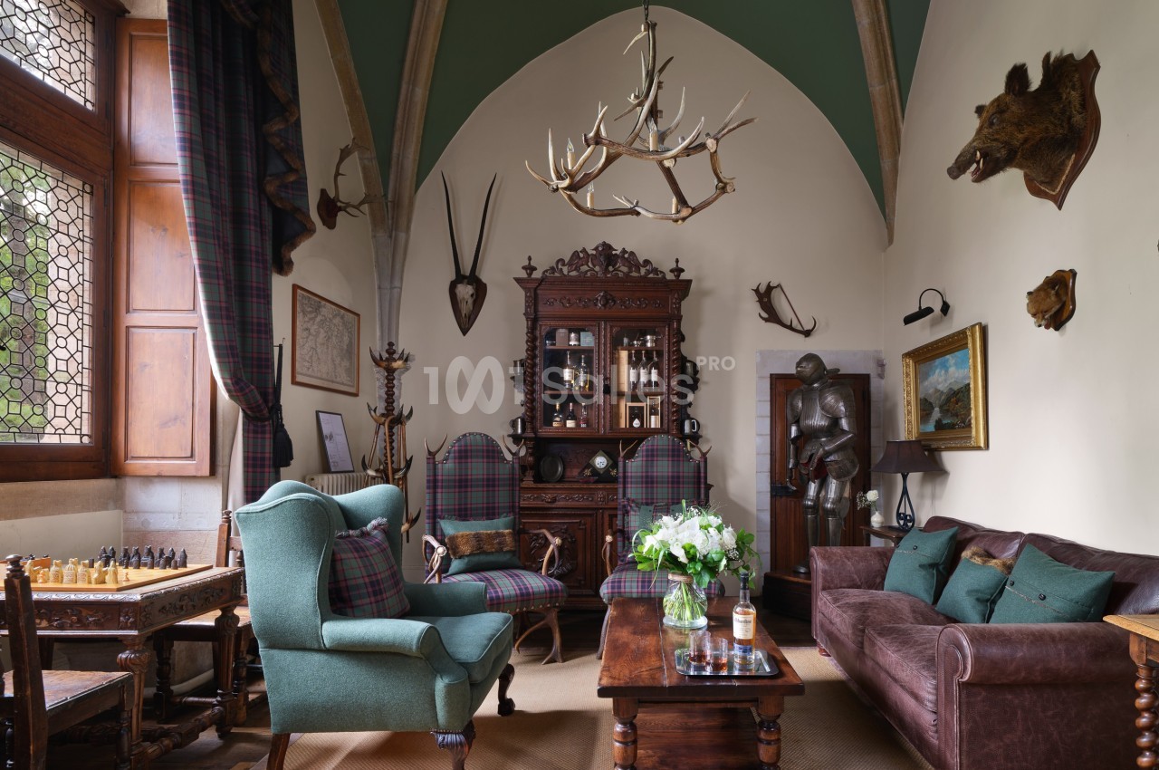 Salon au style médiéval avec mobilier en bois, fauteuils, cheminée, armure, trophées de chasse et plafond voûté.