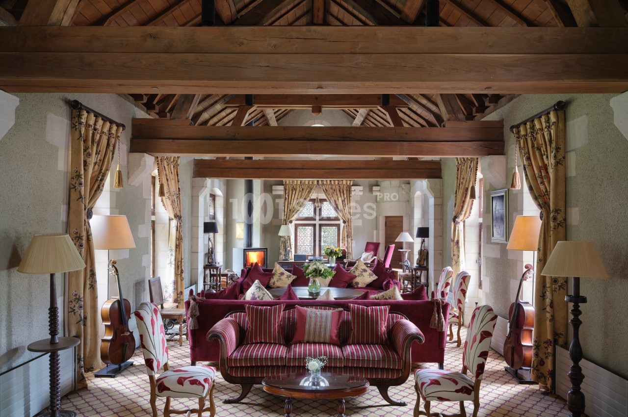 Salon spacieux avec poutres apparentes, mobilier classique, canapés rouges, fauteuils rayés et lumière naturelle.
