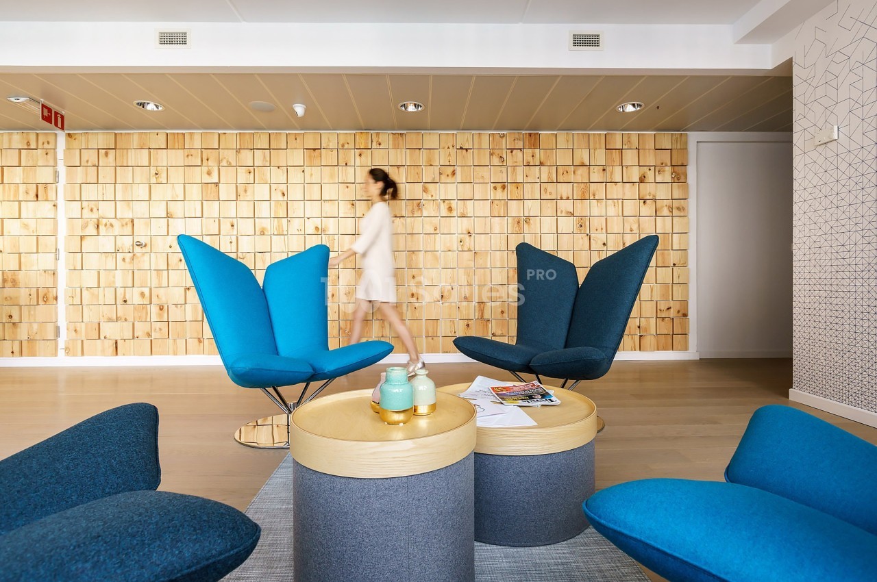 Espace lounge moderne avec fauteuils bleus, tables basses rondes et mur en bois clair, une personne floue en arrière-plan.