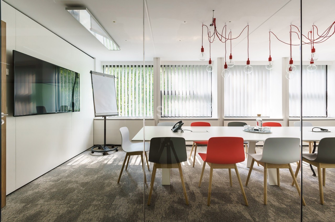 Salle de réunion moderne avec table blanche, chaises colorées, tableau de conférence et grandes fenêtres avec stores.