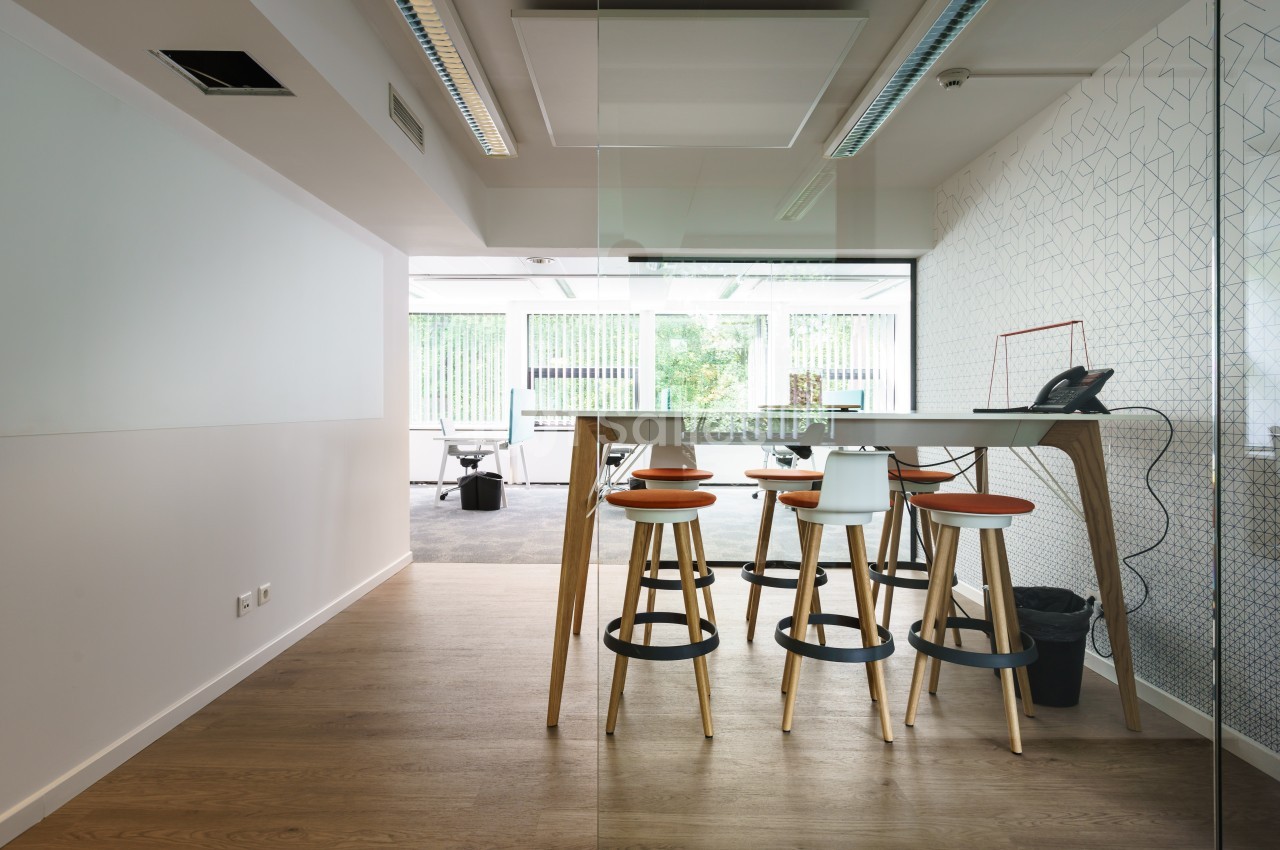 Salle de réunion moderne avec table haute, tabourets en bois et cloison vitrée donnant sur un espace de travail lumineux.