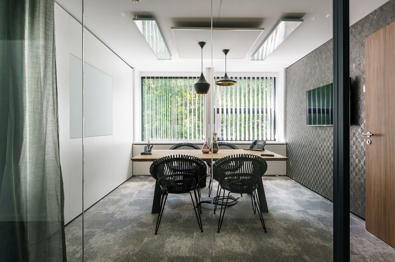 Salle de réunion moderne avec table, chaises noires, luminaires suspendus et grande fenêtre avec stores verticaux.