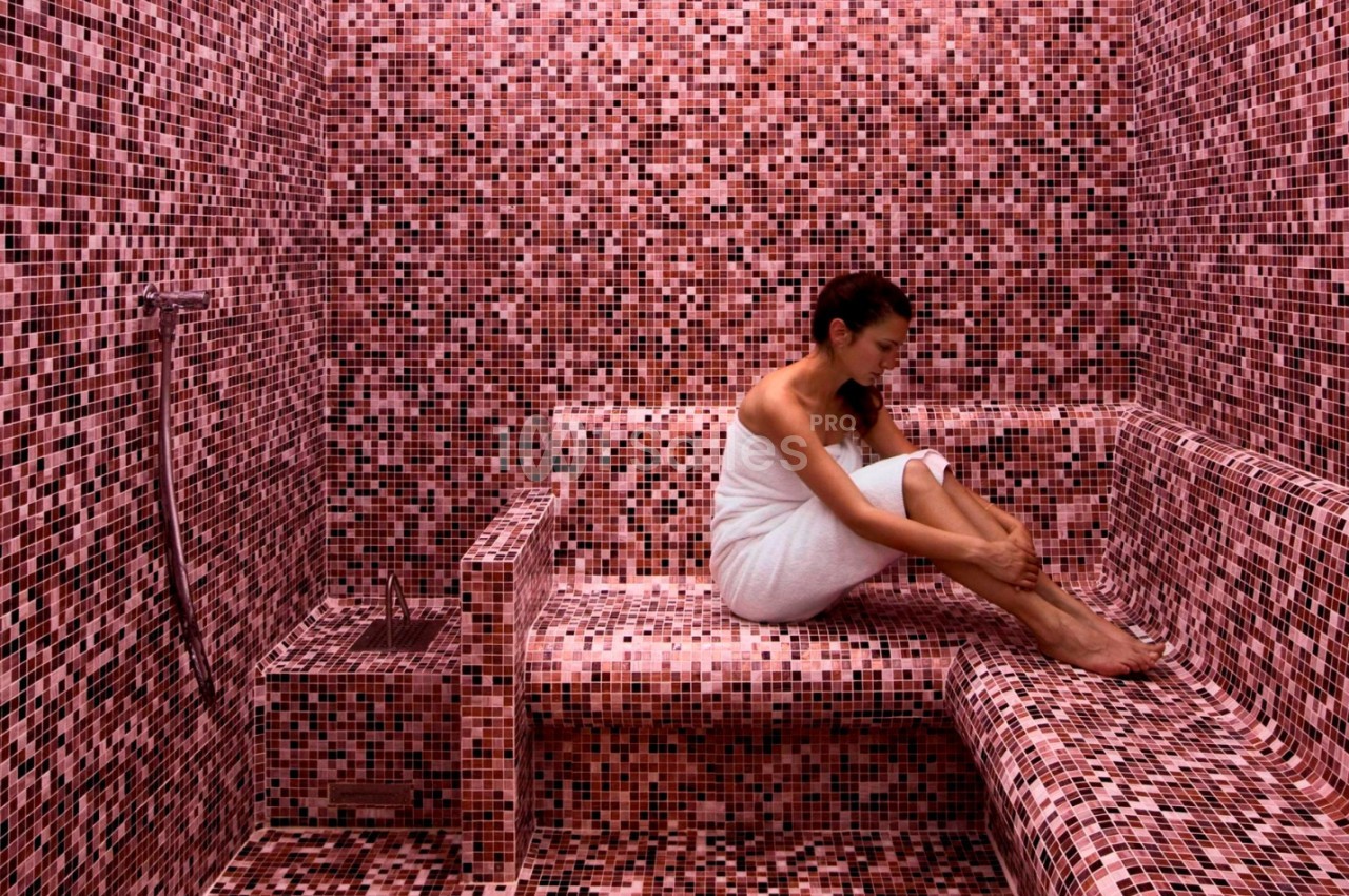 Femme assise dans un hammam aux carreaux roses, enveloppée dans une serviette blanche.