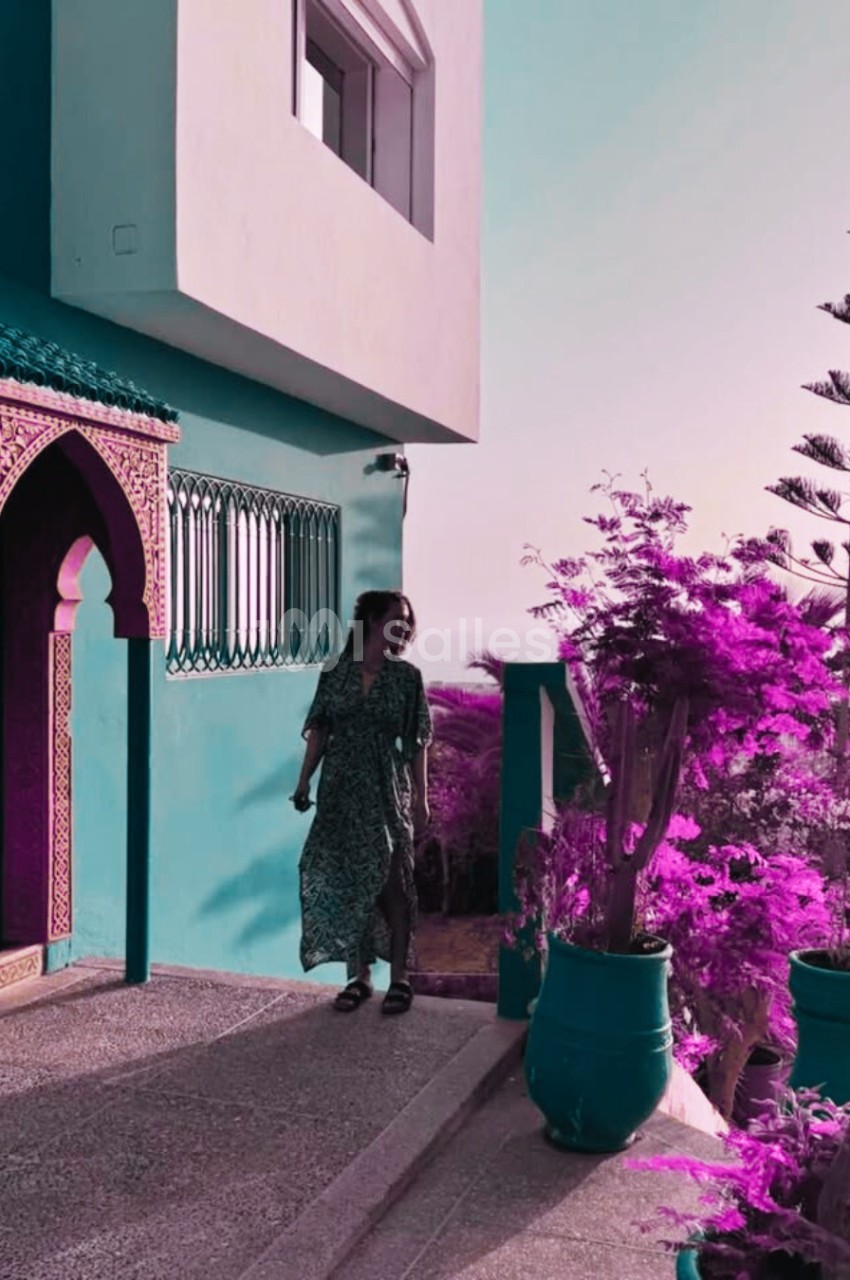 Une femme marche près d'une maison turquoise avec des plantes violettes et un ciel clair en arrière-plan.