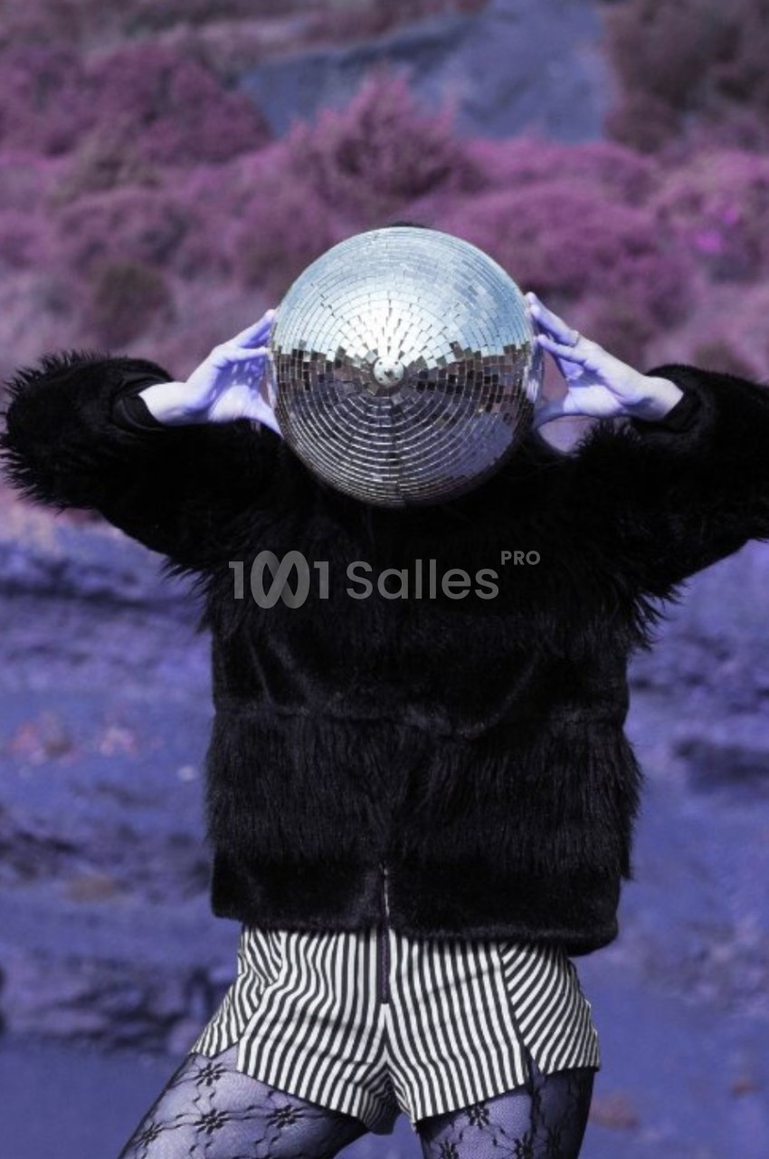 Personne portant une boule à facettes sur la tête, vêtue d'une veste noire et d'un short rayé, dans un paysage violet.