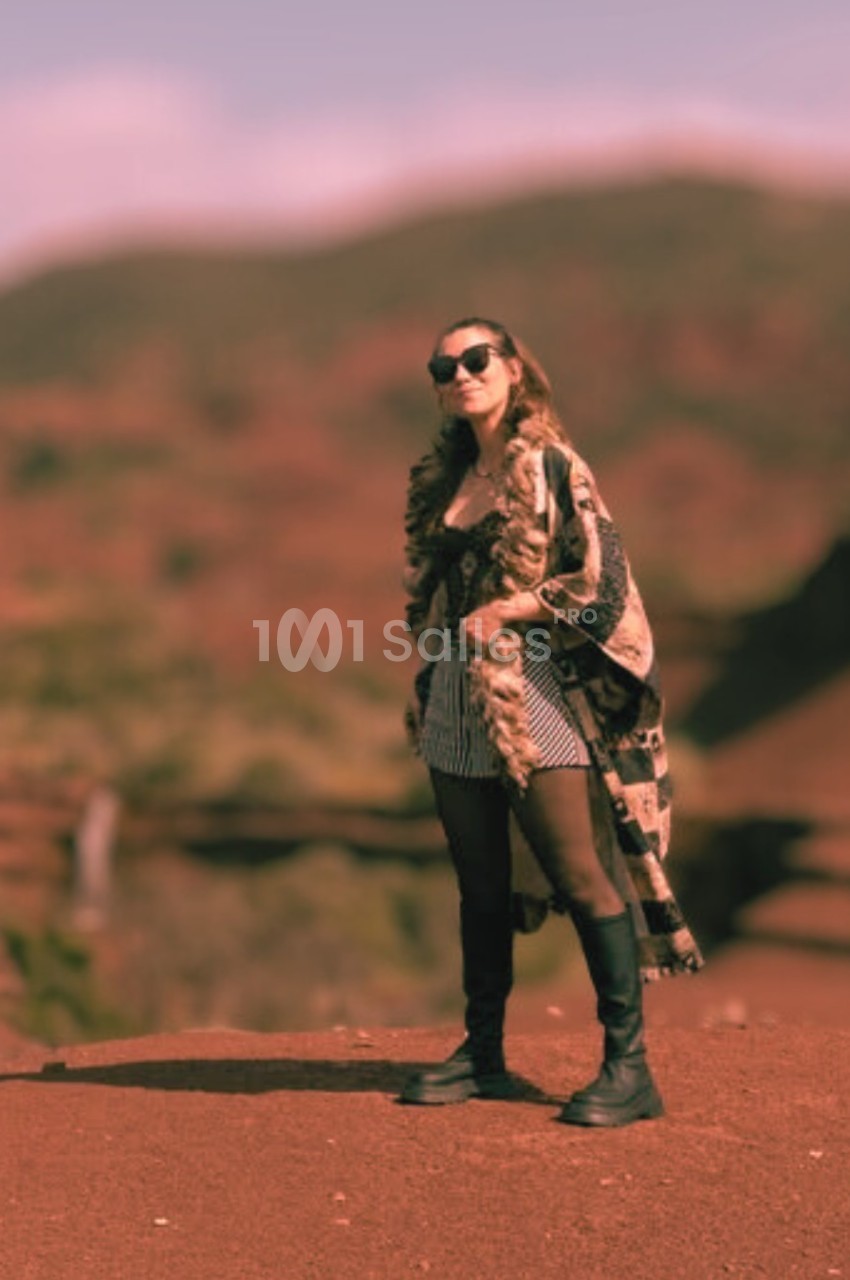 Femme debout sur un sol rouge, portant des lunettes de soleil et un manteau long, avec un paysage flou en arrière-plan.