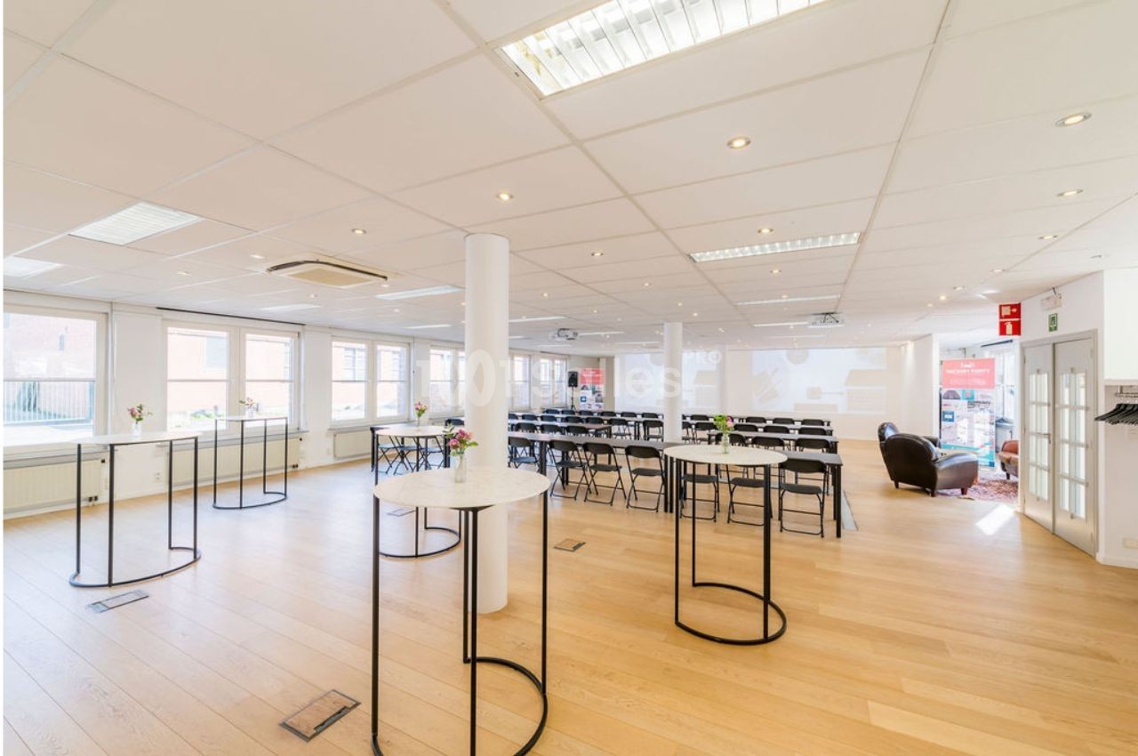 Salle lumineuse avec parquet, tables hautes rondes et rangées de chaises face à un écran de présentation.
