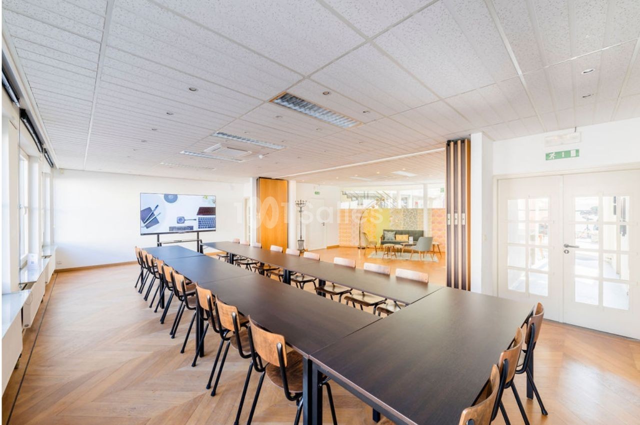 Salle de réunion lumineuse avec de longues tables noires, des chaises en bois et un écran au mur.