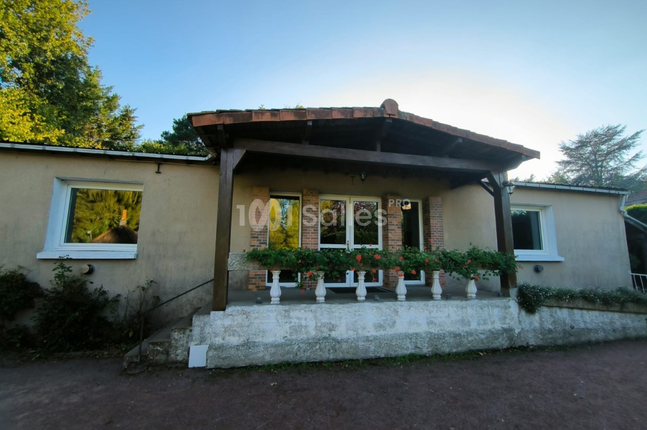 Façade d'une maison avec terrasse couverte, jardinières fleuries et murs clairs, entourée de végétation.