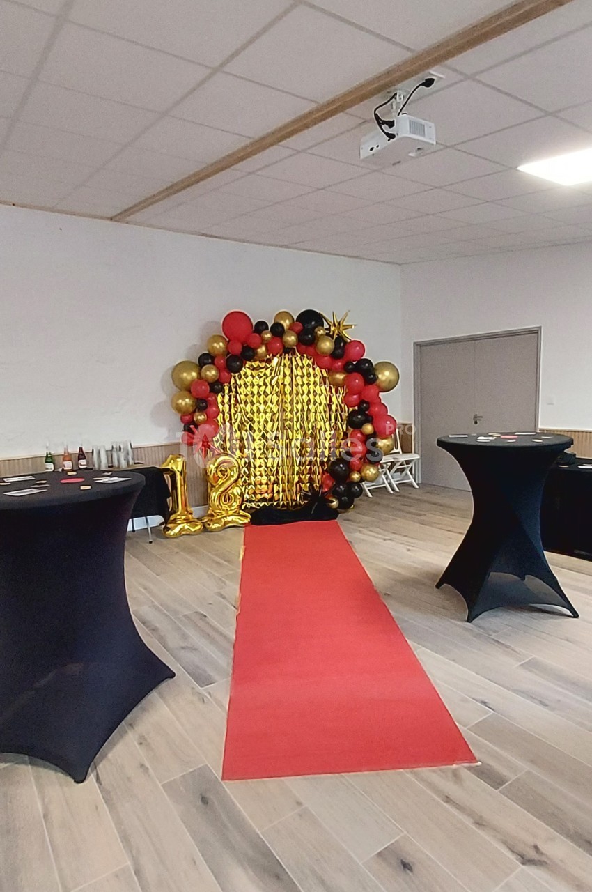Décoration festive avec arche de ballons rouges, noirs et dorés, rideau scintillant et tapis rouge dans une salle.