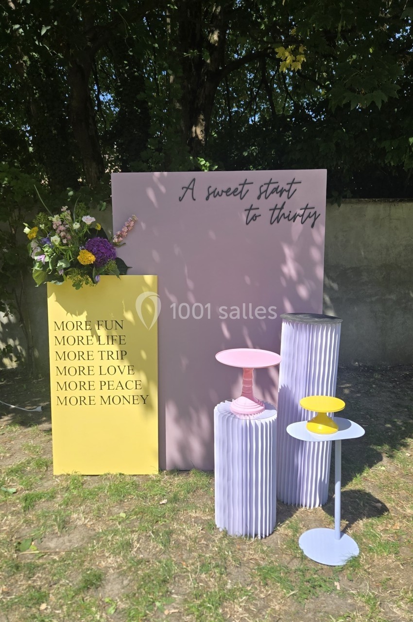 Décor extérieur avec panneaux colorés, fleurs, et présentoirs pastel, sur fond d'herbe et de mur.