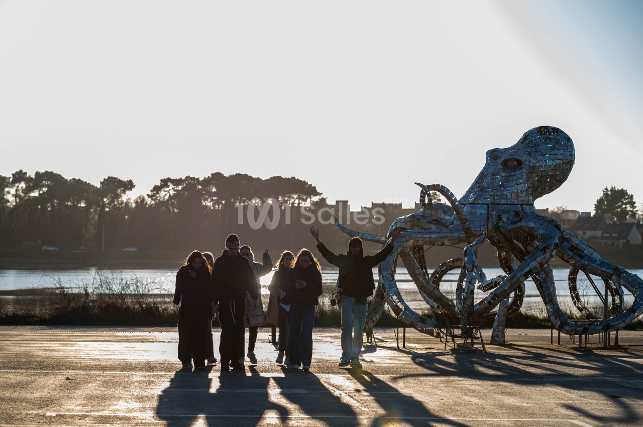 Un groupe de personnes marche près d'une sculpture métallique en forme de pieuvre, avec un paysage naturel en arrière-plan.