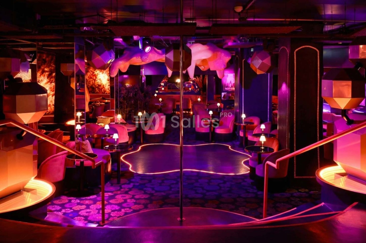 Salle de club avec éclairage néon rose et violet, tables rondes, fauteuils, et décorations géométriques modernes.