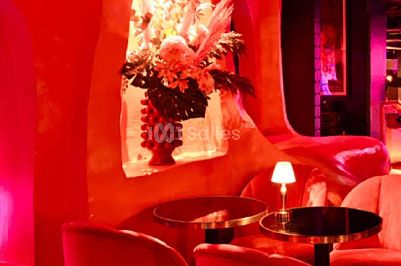Décor intérieur avec fauteuils rouges, tables noires et un vase fleuri éclairé par une lumière tamisée.