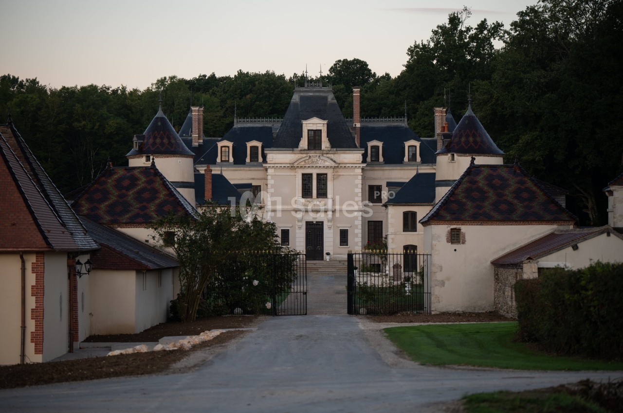 Façade d'un château entouré de dépendances, avec une allée centrale menant à une grille en fer forgé.