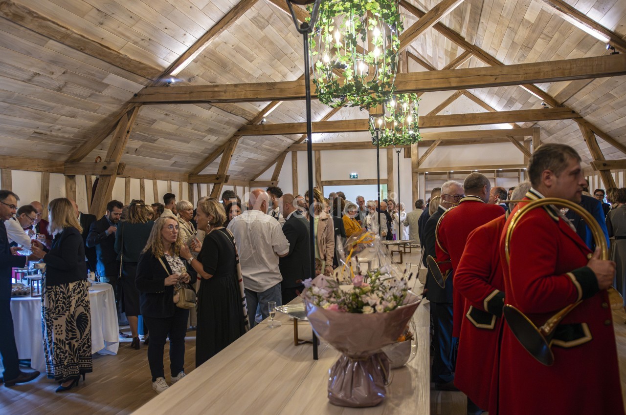 Salle en bois avec des invités en tenue formelle discutant lors d'un événement, décorée de fleurs et de lustres suspendus.