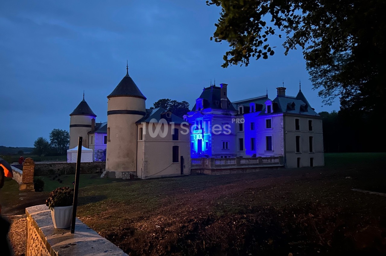 Château éclairé en bleu à la tombée de la nuit, entouré d'arbres et d'un ciel nuageux.