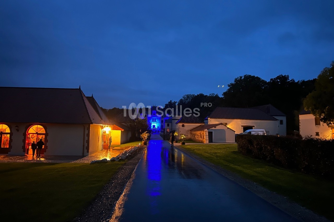 Chemin pavé éclairé menant à un bâtiment illuminé en bleu, entouré de maisons dans un cadre nocturne.