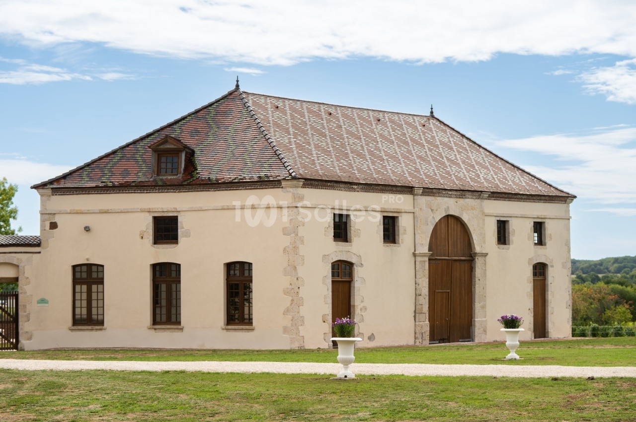Bâtiment ancien avec toit en tuiles, grandes portes en bois et fenêtres, entouré de pelouse et de jardinières fleuries.