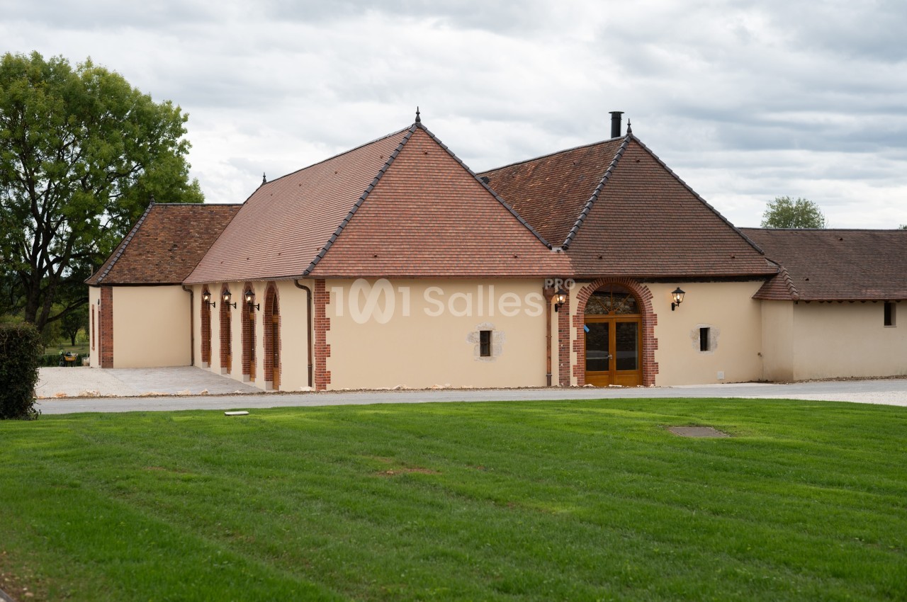 Bâtiment à toit en tuiles rouges avec murs beige clair, entouré de pelouse et d'arbres sous un ciel nuageux.