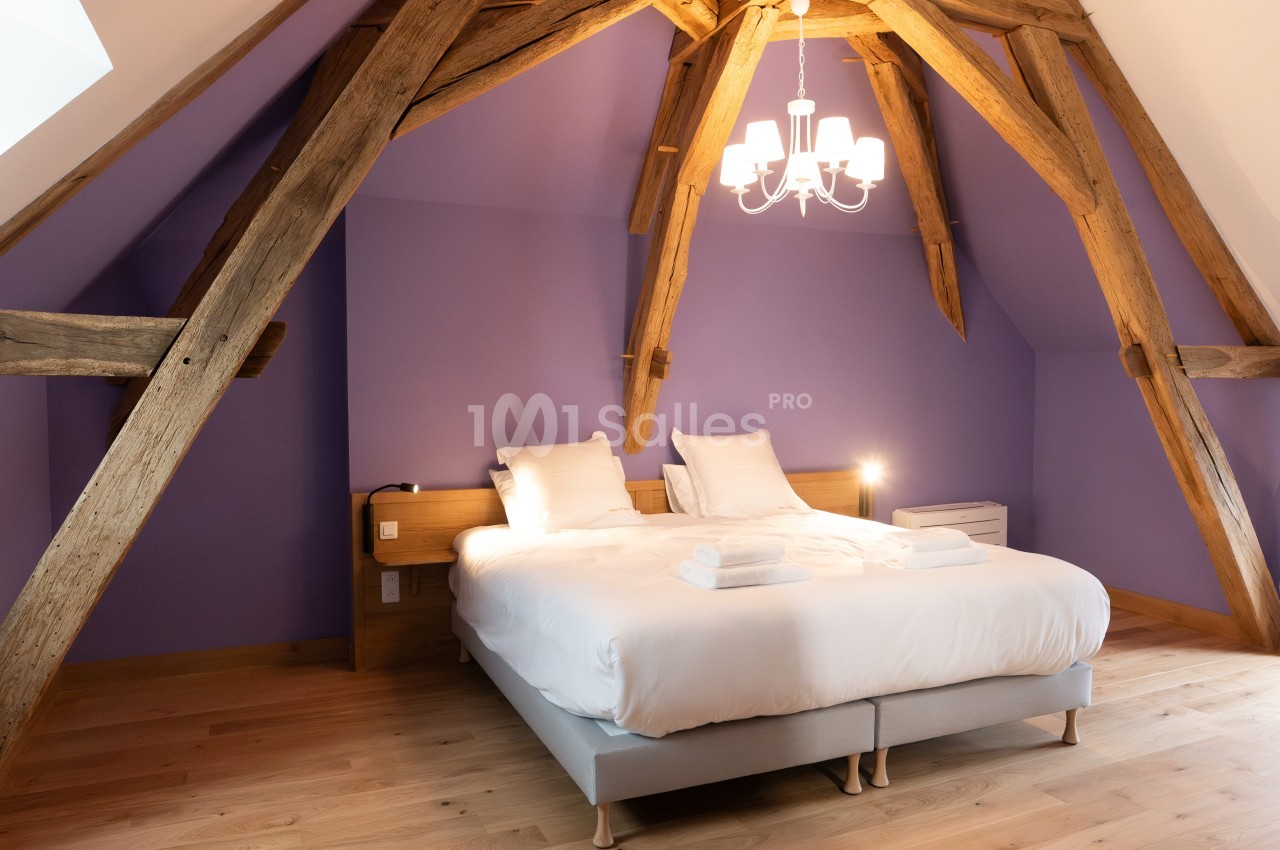 Chambre mansardée avec poutres apparentes, lit double, linge de lit blanc et mur violet, éclairée par un lustre.
