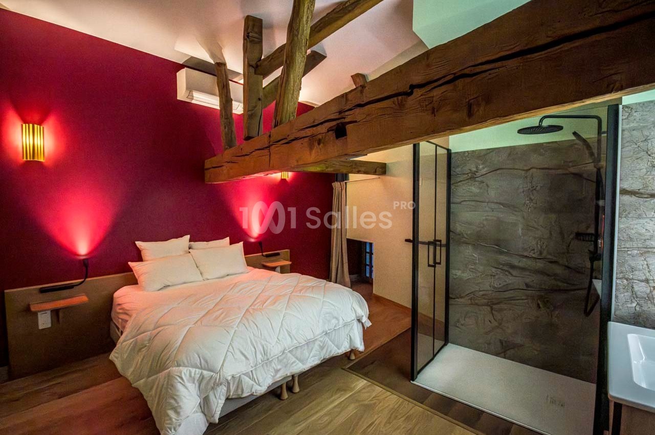 Chambre avec lit double, poutres apparentes, mur rouge, éclairage tamisé et salle de bain ouverte avec douche moderne.