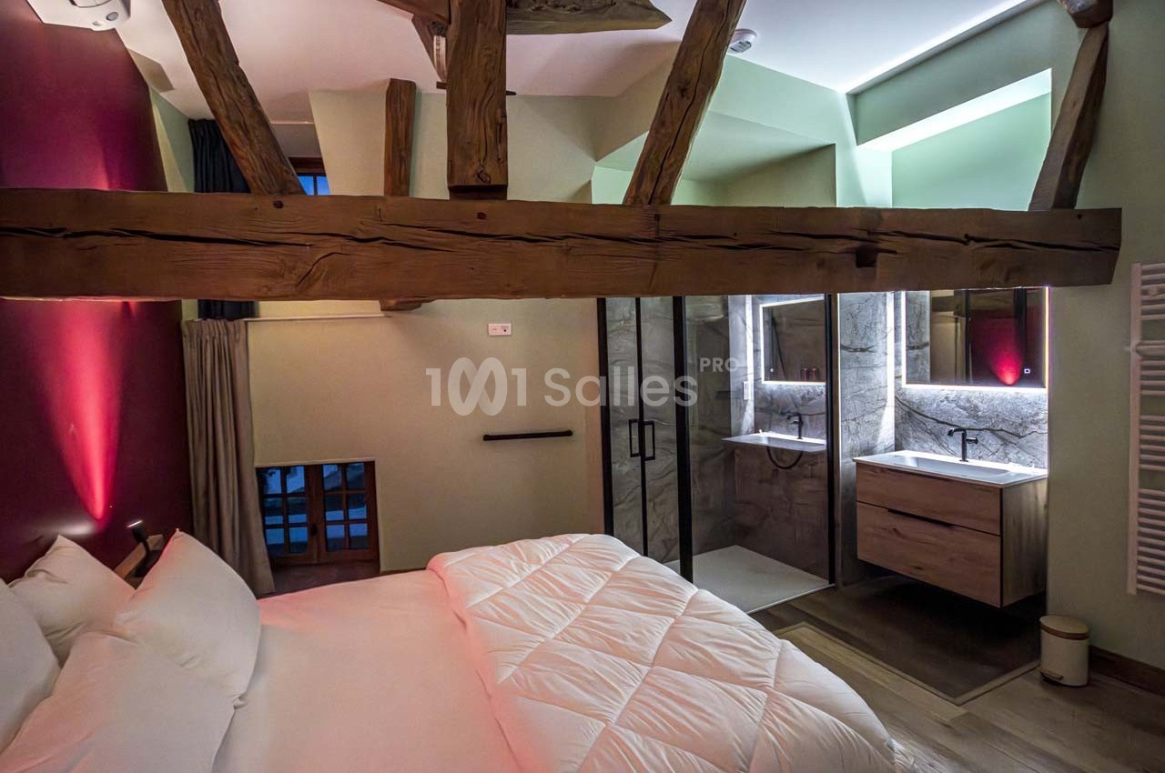 Chambre avec poutres apparentes, lit double et salle de bain moderne attenante avec douche et lavabo éclairés.